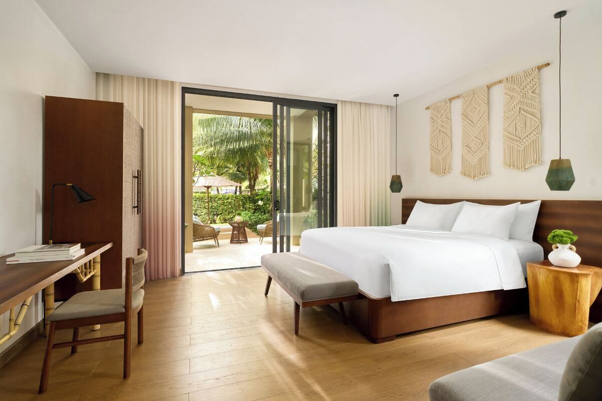 A hotel room with a bed, a desk, and a chair. There is a sliding door leading to an outdoor area with a table and chairs.
