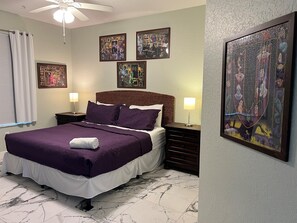 Habitación de hotel con una cama, dos mesitas de noche, un ventilador de techo y obras de arte en las paredes.