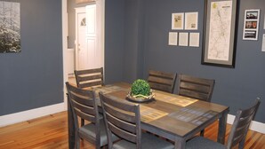 Un coin repas avec une table en bois, six chaises et une petite plante en pot. Des documents encadrés et une carte sont accrochés au mur.