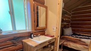 A log cabin bathroom with a wooden vanity, a toilet, and a bed in the adjacent room.
