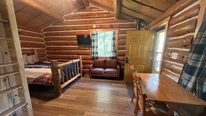 A log cabin interior with a wooden floor, a sofa, a dining table, and a bed with a wooden headboard.