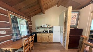 A log cabin interior with a wooden dining table, chairs, a kitchen area with wooden cabinets, a refrigerator, and a window with curtains.