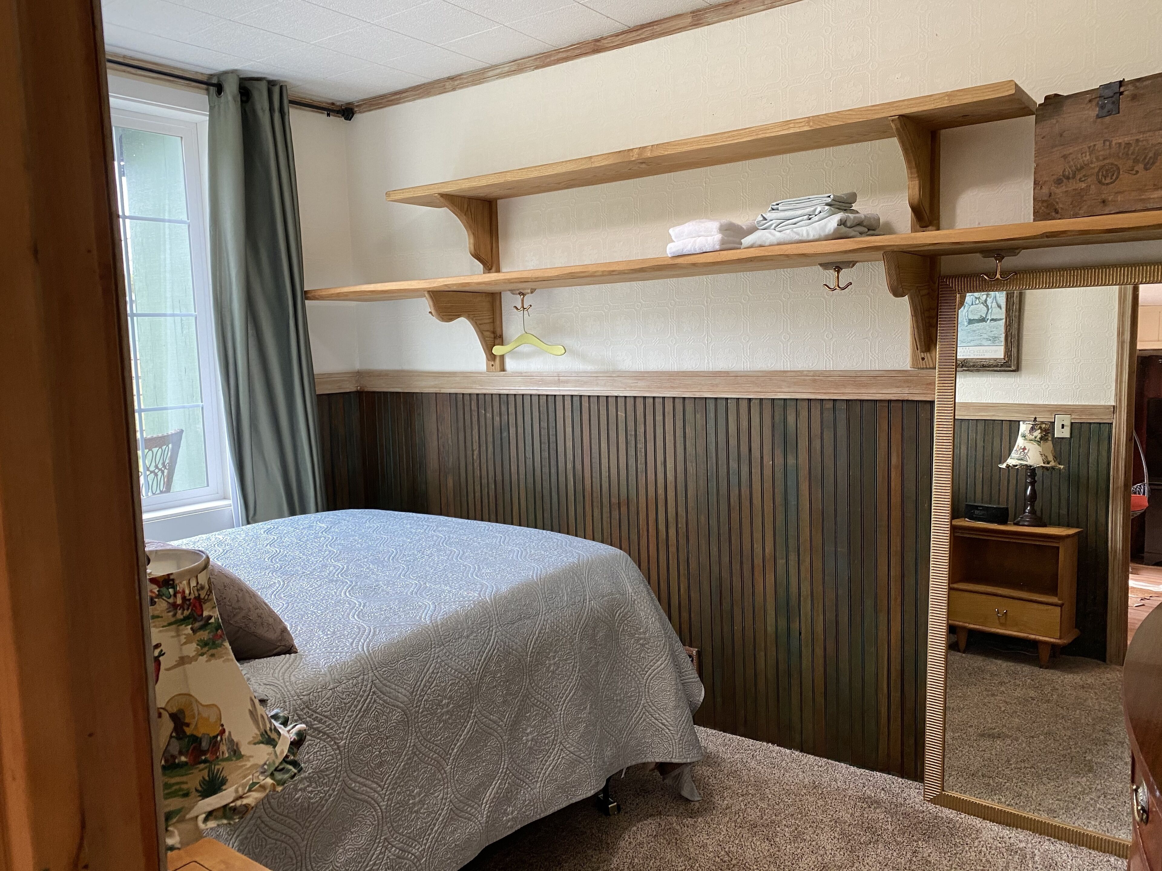 Ein Schlafzimmer mit einem Bett, einem Holzbodenregal, einem Spiegel und einem Fenster mit Vorhängen.