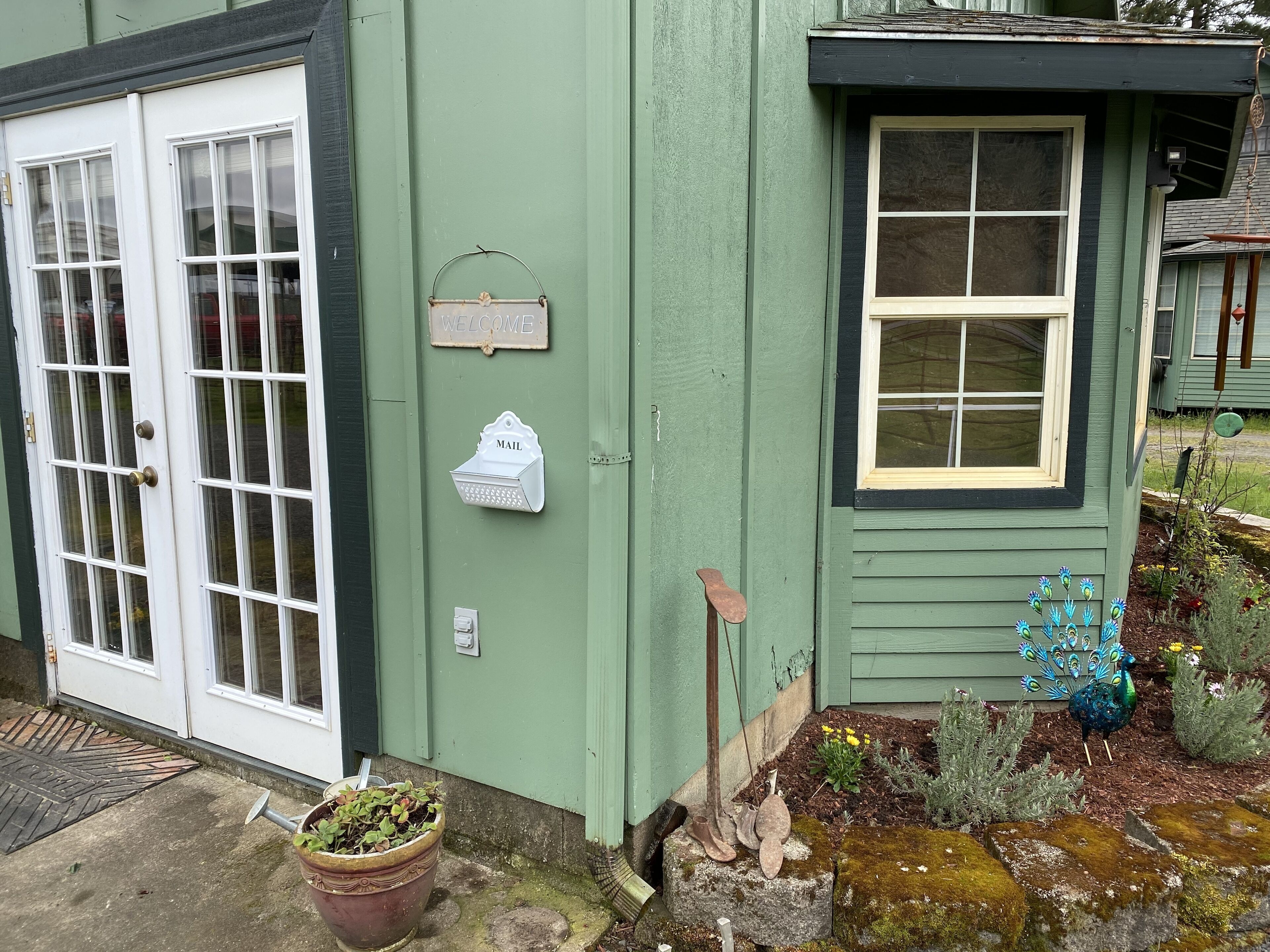 A green building with a white door and windows, a mailbox, and a small garden with peacock statues.