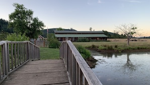 Un pont en bois mène à un bâtiment rustique surmonté d’un toit vert, entouré de verdure et bordé par une étendue d’eau calme.