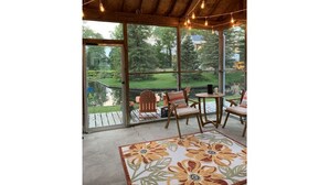 Eine überdachte Terrasse mit großen Fenstern, Holzmöbeln und einem floralen Teppich.