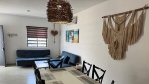 A modern living room with a sofa, dining table, chairs, a hanging macrame wall decoration, and a painting on the wall.