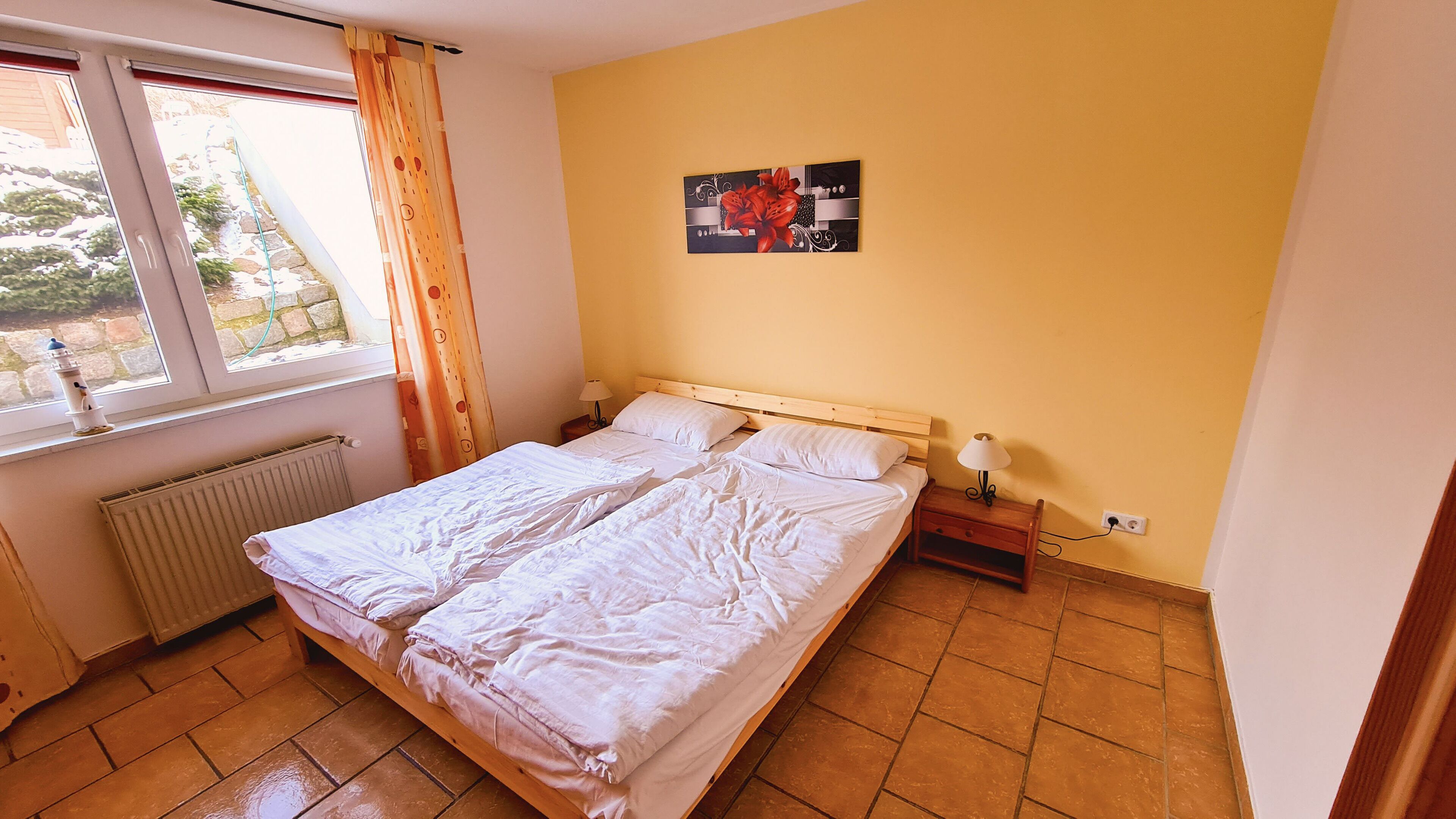 A bedroom with a large bed, a window with curtains, a radiator, a nightstand with a lamp, and a wall-mounted picture.