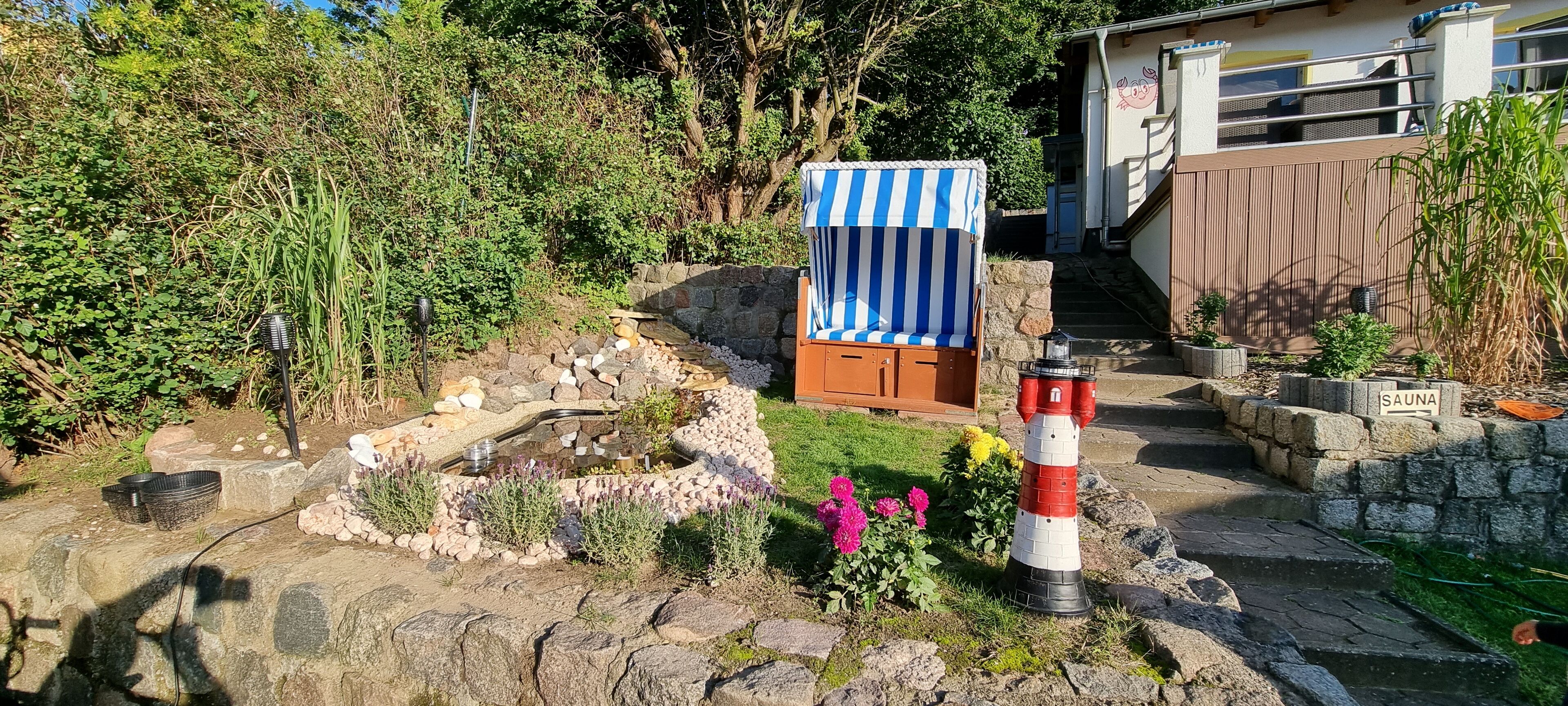 Un giardino con una piccola piscina, una panchina di legno, un cartello che indica una sauna e un modellino di un faro rosso e bianco.