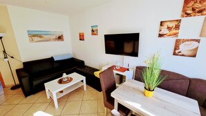 Ein Wohnzimmer mit einer schwarzen Couch, einem weißen Couchtisch, einem braunen Sessel, einem Fernseher und einem kleinen Tisch mit einer Pflanze.