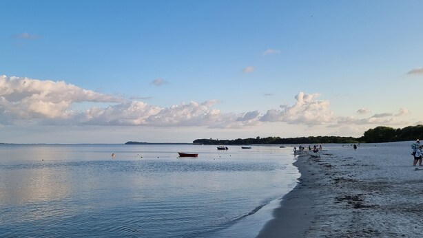Ein Strand mit ruhigem Wasser, Booten und Menschen am Ufer.