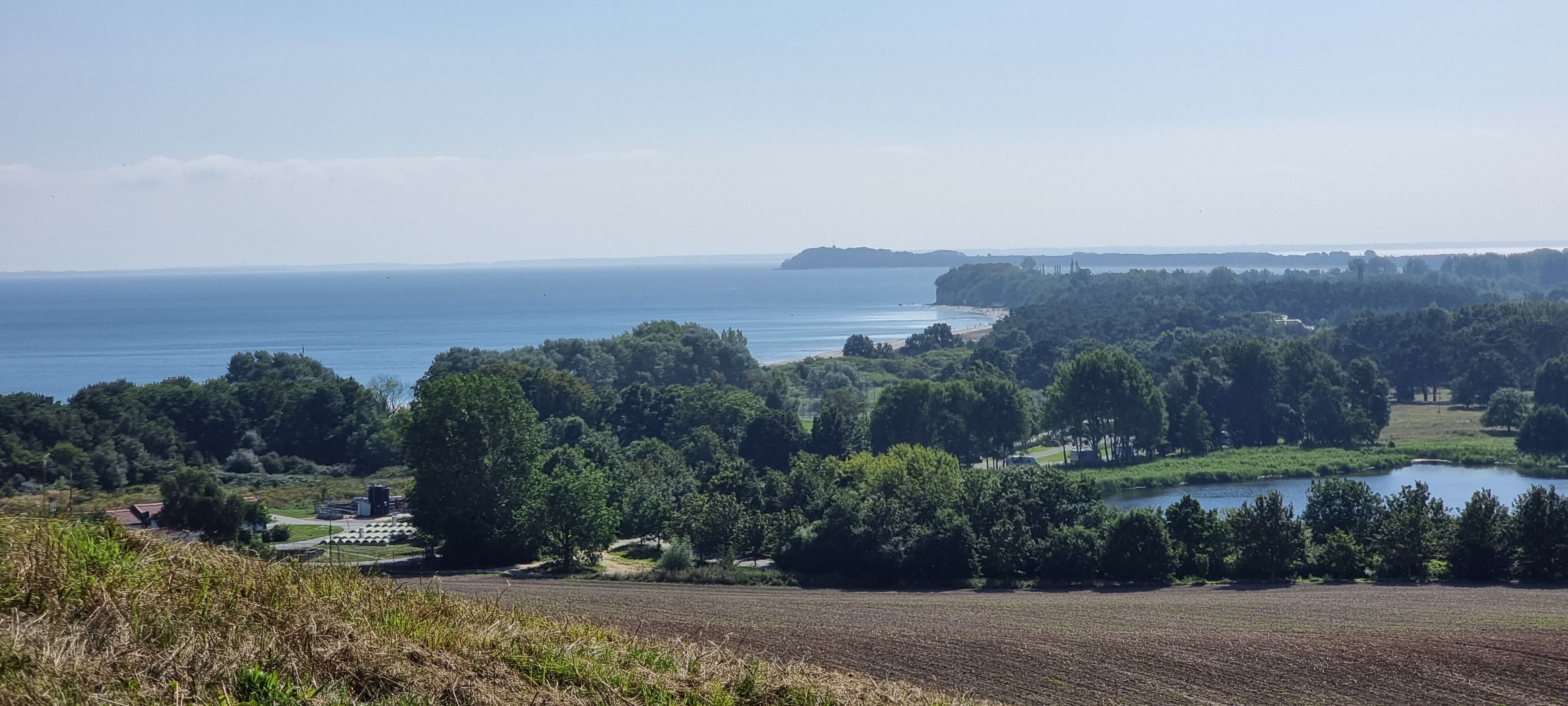 A coastal landscape with a large body of water, dense greenery, and a few buildings in the distance.