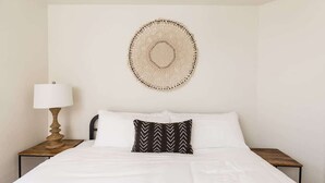 A bed with white bedding, a black and white patterned pillow, a wall-mounted circular woven artwork, and a bedside lamp on a wooden nightstand. - Western Hills-Pool Heat Avail- Putting Green-Games (Fountain Hills)