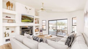 A modern living room with a sofa, TV, fireplace, and a view of the outdoors. - Western Hills-Pool Heat Avail- Putting Green-Games (Fountain Hills)