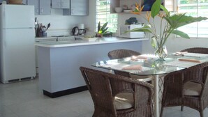 Une cuisine avec des meubles blancs, un réfrigérateur blanc, une machine à café et un coin repas avec des chaises en osier et une table en verre.
