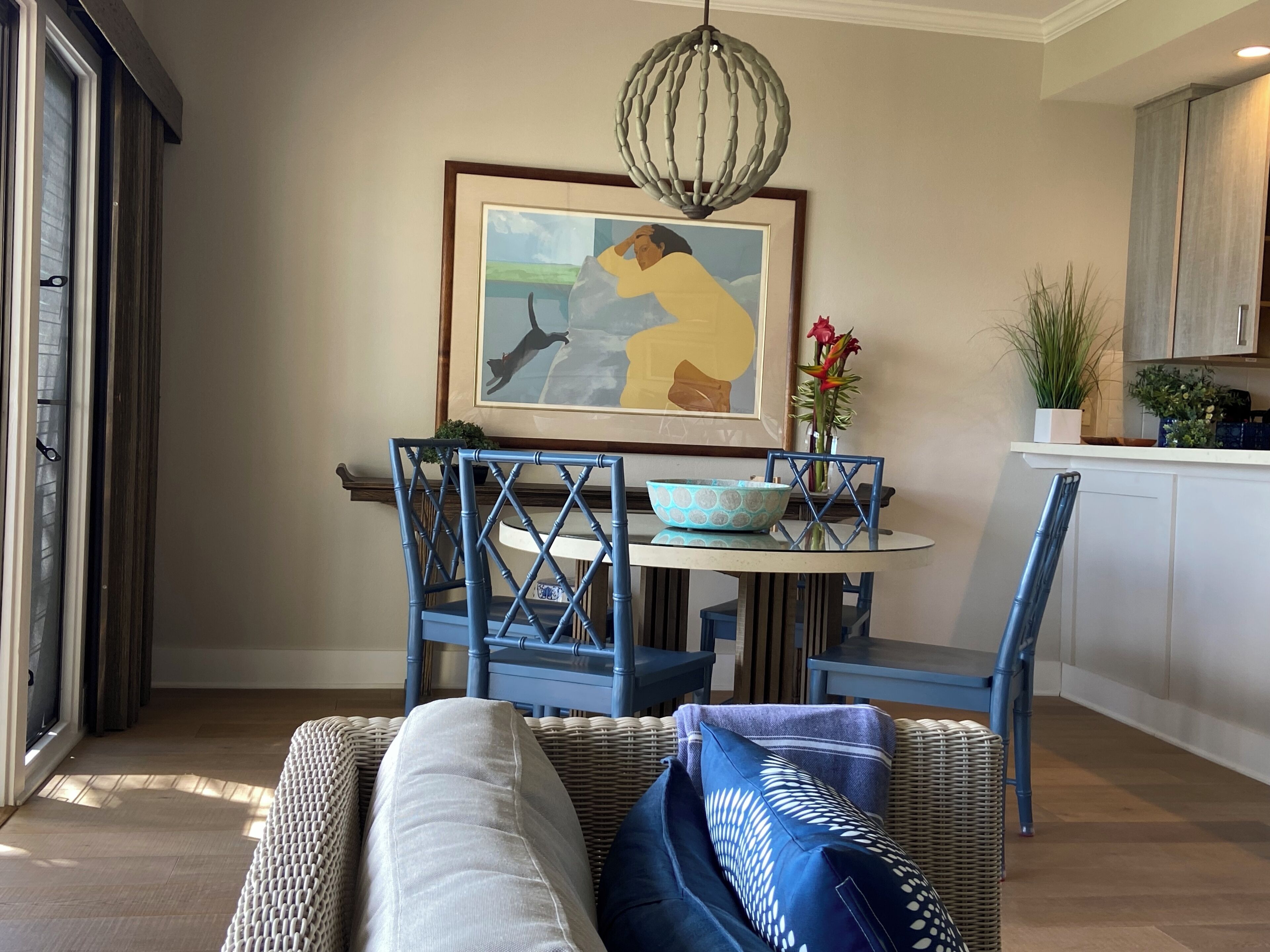 A dining area with a round table, blue chairs, a framed painting, and a vase with flowers.