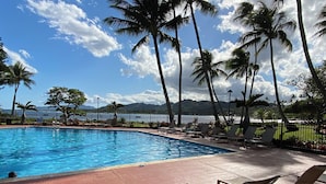 A swimming pool with lounge chairs and palm trees, with a view of the ocean and mountains in the background.