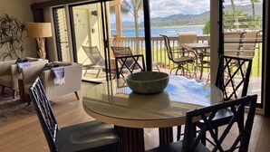 A modern living room with a glass table and chairs, a sofa, and a view of the ocean and hills.
