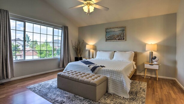 A bedroom with a large window, a ceiling fan, a bed with white bedding, a bedside table with a lamp, and a small nightstand with a fan. - Spacious Thornton Home: 15 Mi to Downtown Denver! (Thornton)
