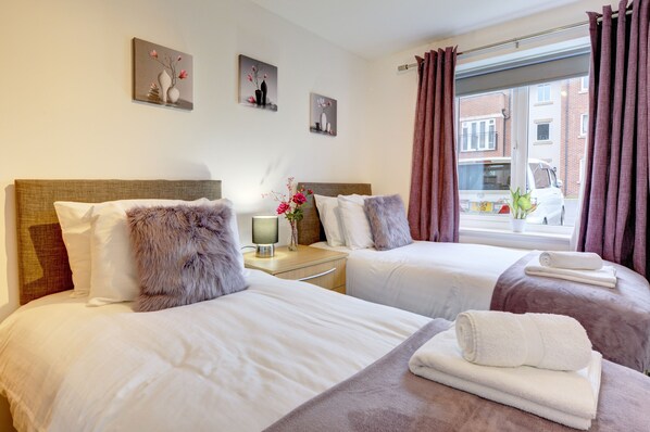 A hotel room with two beds, a nightstand with a lamp, and a window with curtains.