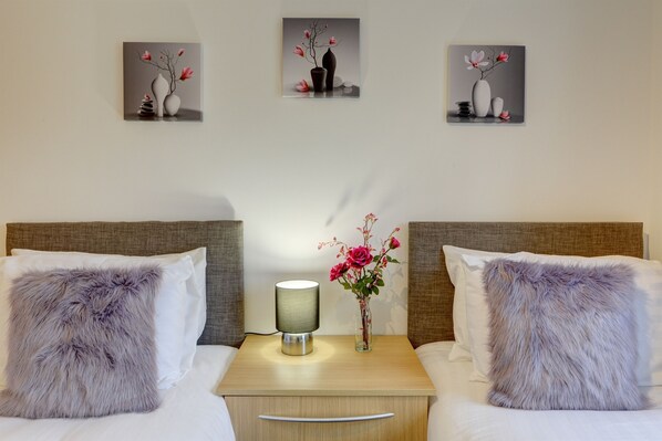 Een hotelkamer met twee bedden, een nachtkastje met een lamp, een vaas met bloemen en drie ingelijste kunstwerken aan de muur.