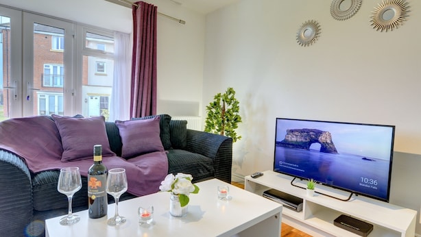 Una sala de estar moderna con un sofá, una mesa de centro con copas de vino y una botella, un televisor sobre un mueble bajo y decoración en las paredes.