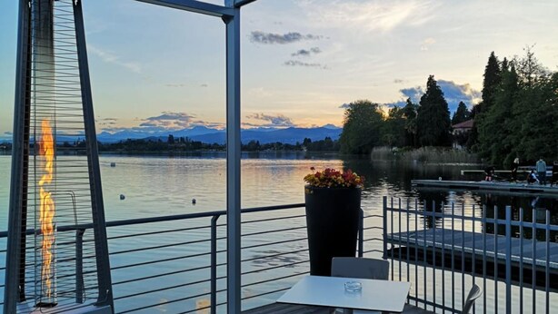 Eine Seeterrasse mit Tisch und Stühlen, mit Blick auf einen stillen See und ferne Berge.