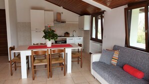 Eine moderne Küche mit weißen Schränken, einem Esstisch mit Stühlen, einer roten Tischmatte und einem Blumentopf. Es gibt ein graues Sofa mit einem roten Kissen und einem gestreiften Kopfkissen.