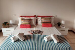 Un lit double avec une literie à rayures, deux oreillers rouges et deux oreillers blancs. Il y a deux tables de chevet avec des lampes et une petite plante sur chacune. Un tapis à rayures est posé sur le sol, et deux serviettes sont pliées sur le lit.