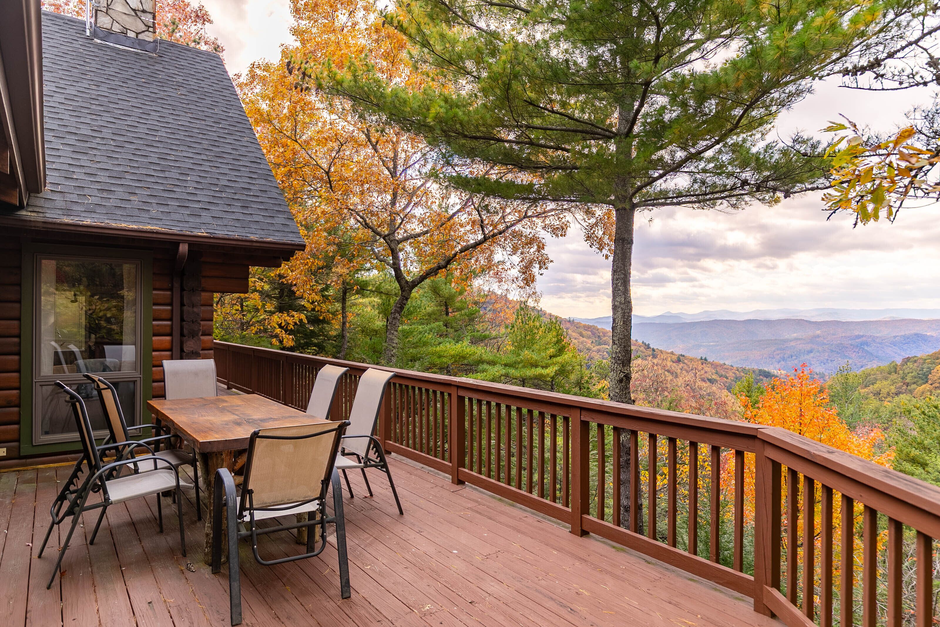 A wooden deck with a table and chairs, overlooking a forested landscape with autumn foliage.
