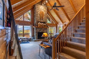 A log cabin interior with a stone fireplace, wooden staircase, and large windows offering a view of the outdoors.