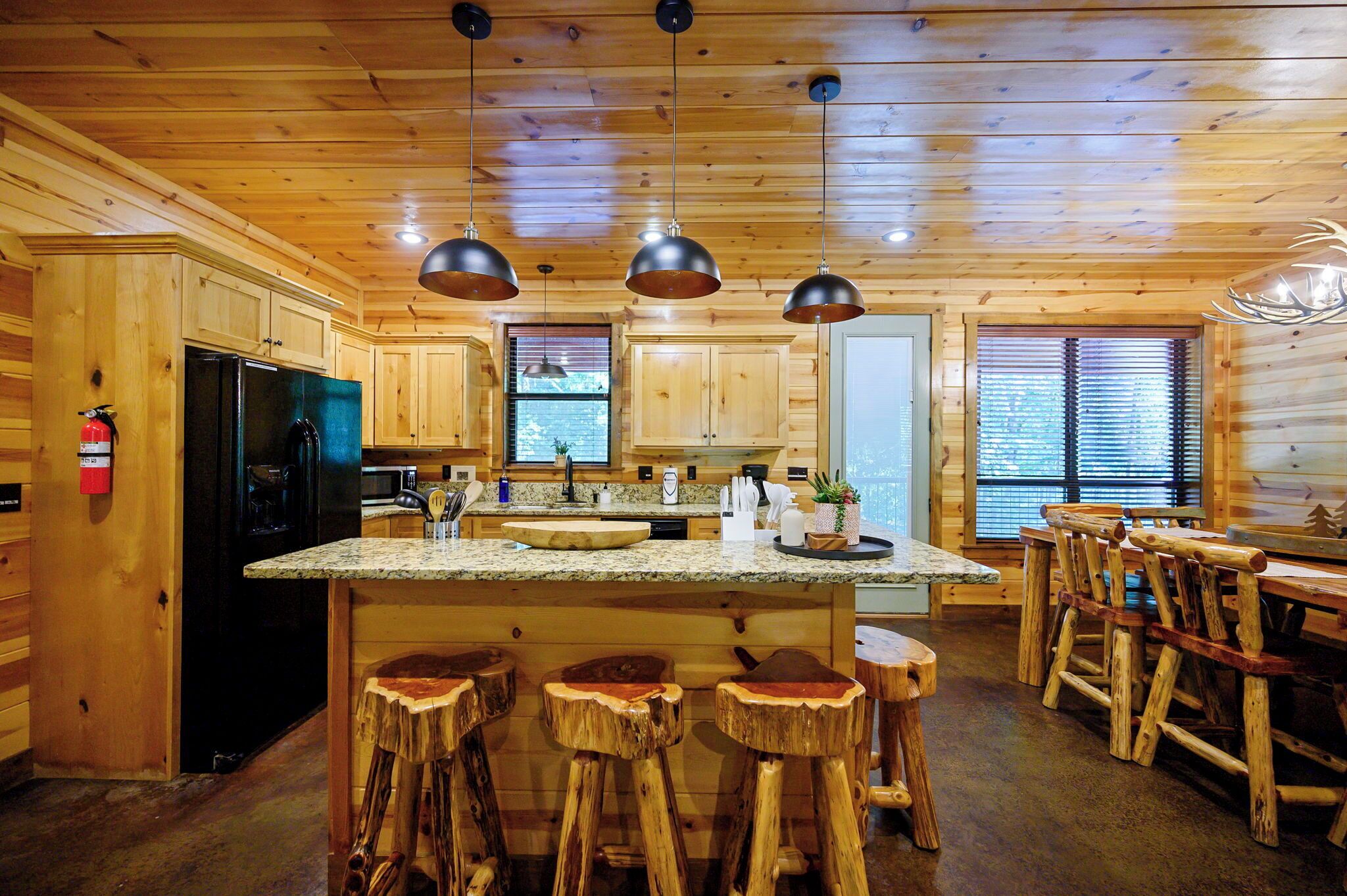 Intérieur d’une cabane en bois avec un espace cuisine comprenant un îlot central, des tabourets de bar et des luminaires suspendus.