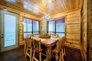Un interior de madera con una mesa redonda de comedor y sillas, una araña y ventanas amplias con persianas.