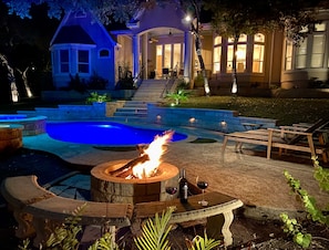 Piscina con pozo de fuego y sillones de descanso, rodeada por un jardín ajardinado y una casa con porche.