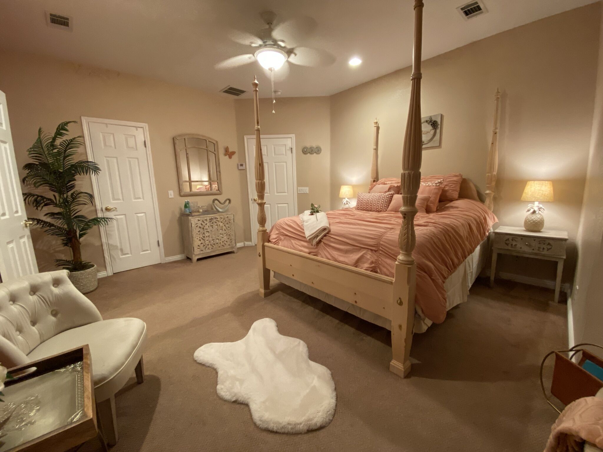 Une chambre à coucher avec un lit à baldaquin, un tapis blanc et moelleux, une chaise, une commode, un miroir et un ventilateur de plafond.