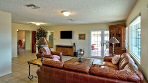A spacious living room with a brown leather sofa, a coffee table, and a television mounted on the wall. There is a dining area visible through a glass door.
