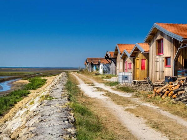 Une rangée de cabanes en bois avec des toits rouges, un chemin de terre menant à ces cabanes, et une vue sur la mer et le paysage plat en arrière-plan.