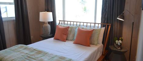Une chambre à coucher avec un lit, une table de chevet avec une lampe, une petite table avec une plante et une fenêtre avec des rideaux.