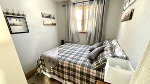 Un dormitori amb un llençol escocès, una taula de nit amb una làmpada i una finestra amb cortines.