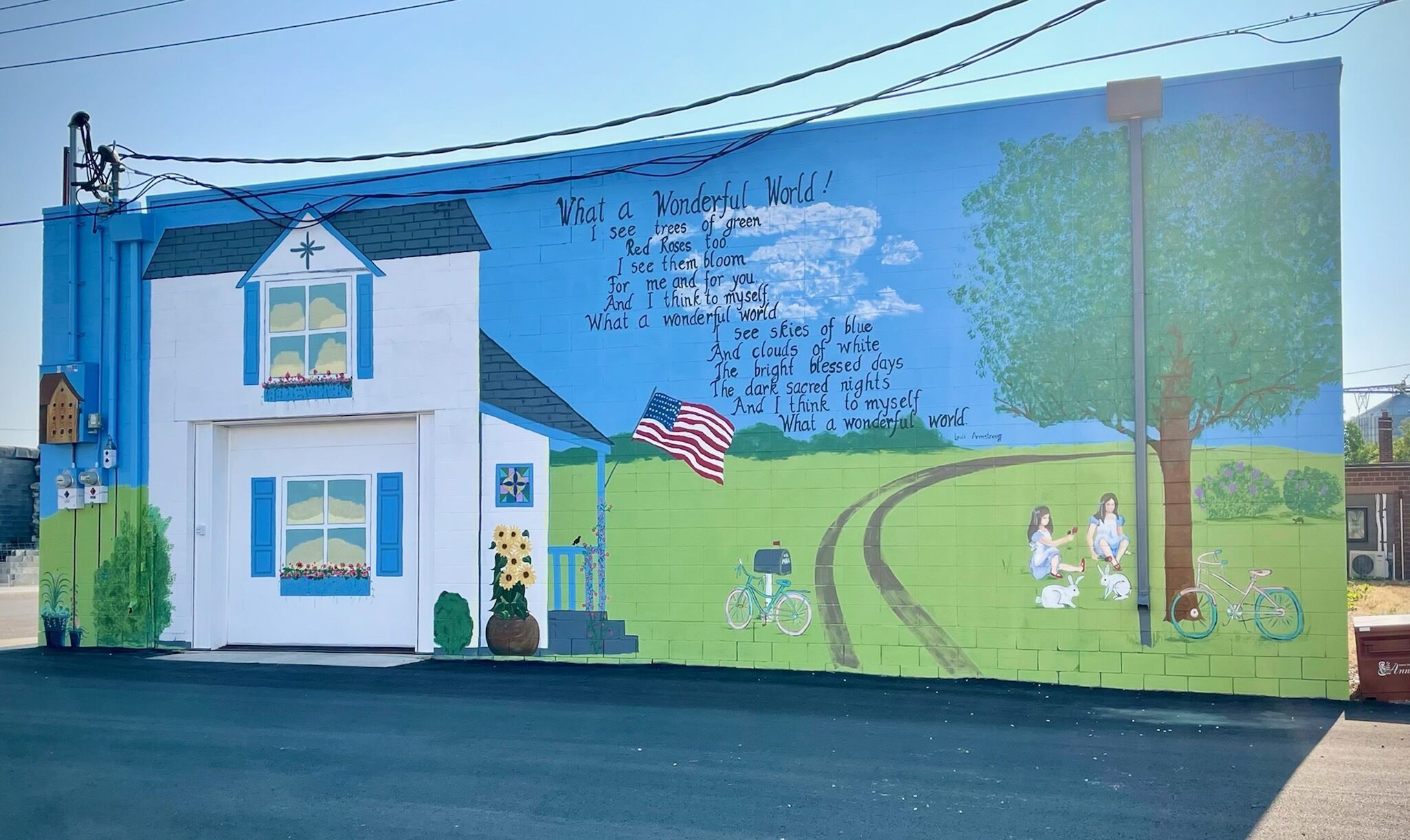 Une fresque murale orne un bâtiment et représente une maison, un arbre et un vélo, accompagnée d’une citation sur le monde.