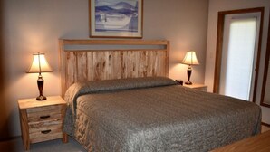 Un dormitorio con cabecera de madera, dos mesitas de noche con lámparas, una cama con colcha estampada, una mesita de noche con cajones y un cuadro enmarcado en la pared.