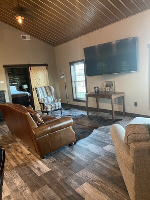 A spacious living room with a large flat-screen TV mounted on the wall, leather sofas, a wooden ceiling, and a large window providing natural light.