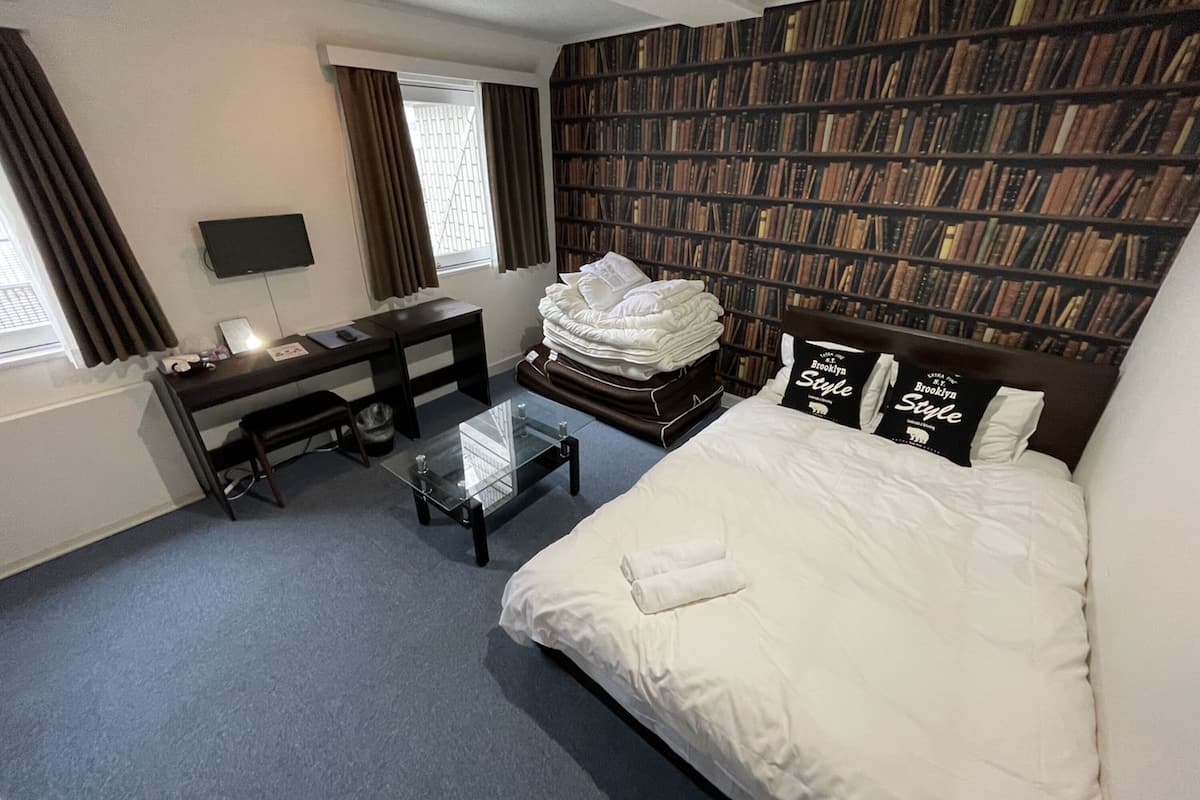 A hotel room with a large bed, a desk with a chair, a glass coffee table, a bookshelf, and a TV on the wall.