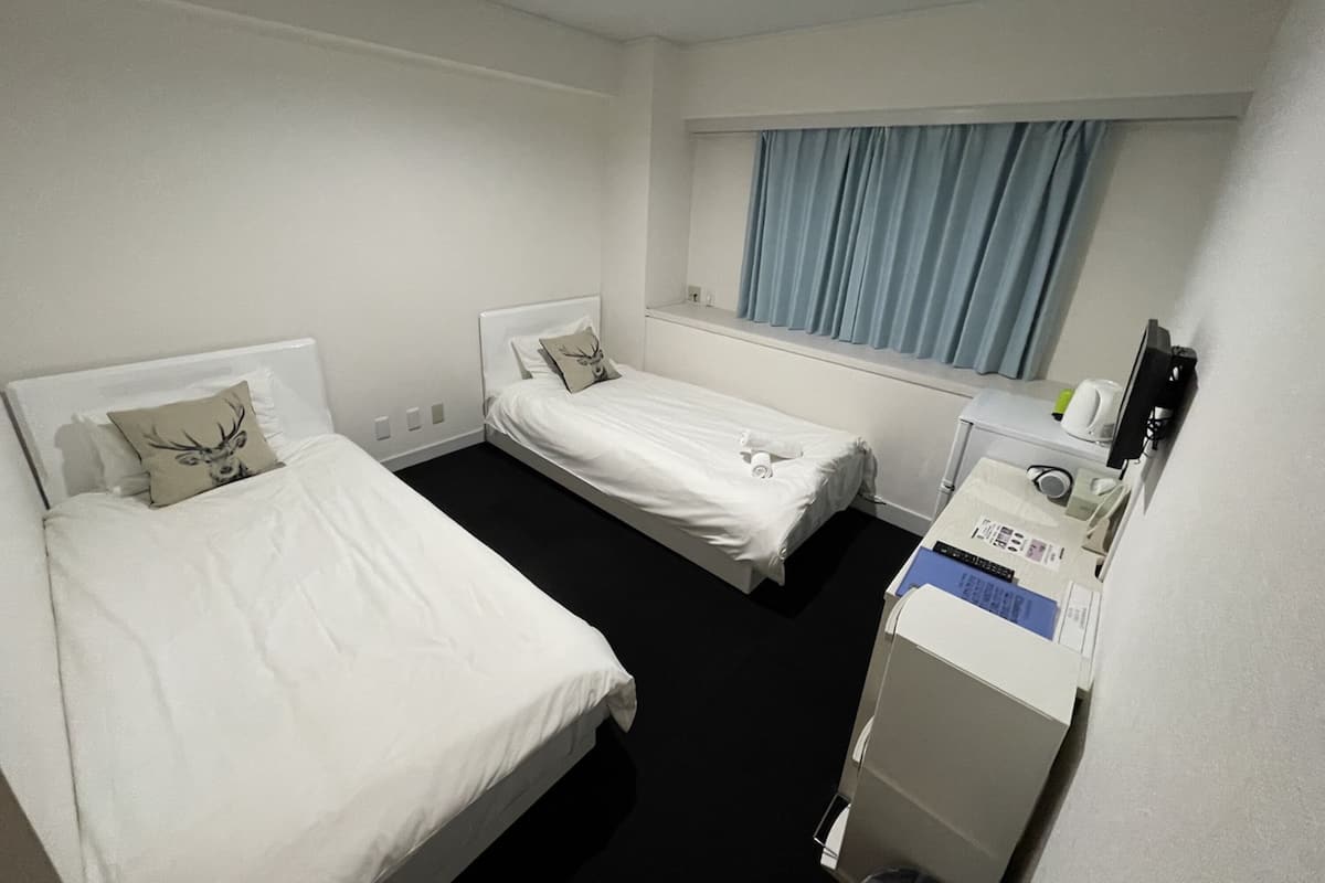 A hotel room with two single beds, a small desk with a printer, and a window with curtains.