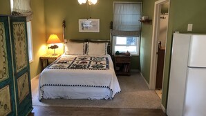 A bedroom with a bed, a nightstand, a lamp, a ceiling fan, a window with blinds, and a refrigerator. - Charming guest house on 1.5 acre lake front property (Green Lake)