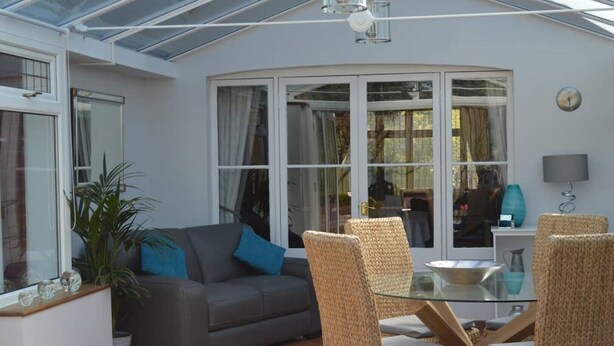 A sunlit room with a glass table, wicker chairs, a grey sofa, and a large window with curtains. - Large four bedroom home near country walks & beach (Shanklin)
