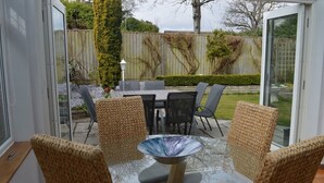 A patio area with a glass table surrounded by wicker chairs, a dining table with chairs, and a view of a backyard with a wooden fence and greenery. - Large four bedroom home near country walks & beach (Shanklin)