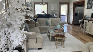 A spacious living room with a large white artificial tree, a beige sofa, a coffee table, and a dining area with a chandelier. - Cabin with Spa and Arcades 7 mins to Ski Resort (Running Springs)