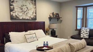 A hotel room with a large floral artwork, a bed with white bedding, a bedside table with a lamp, a shelf with decorative items, and a window with blinds. - Cabin with Spa and Arcades 7 mins to Ski Resort (Running Springs)