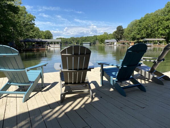 Eine Holzterrasse mit zwei Adirondack-Stühlen, die auf einen ruhigen See ausgerichtet sind, umgeben von Bäumen und Booten, die am Ufer liegen.